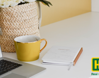 a yellow mug and notebook sit by a laptop on a desk as a howard hanna agent trains offscreen