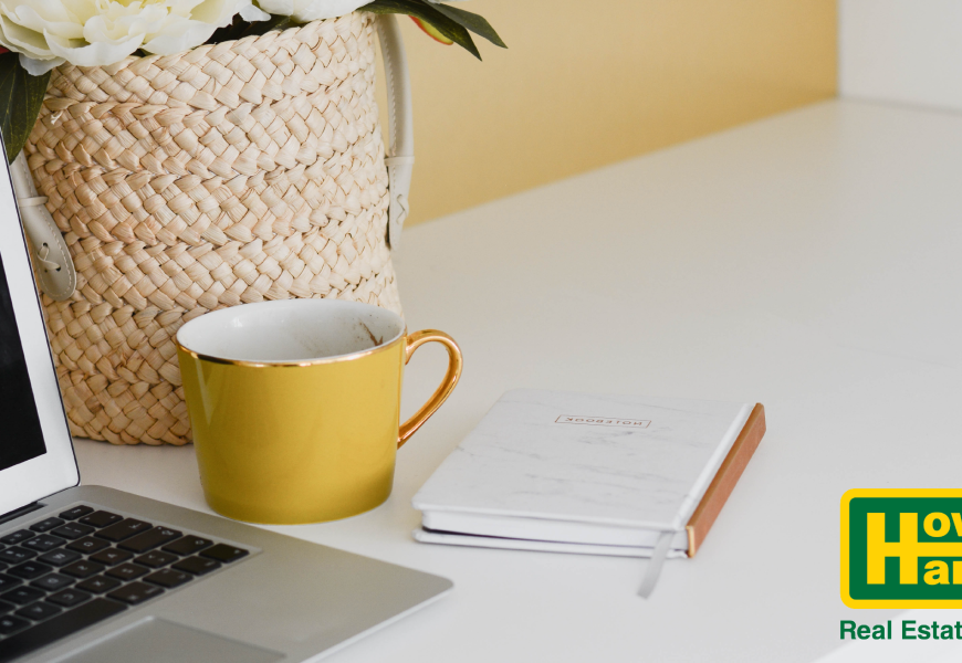 a yellow mug and notebook sit by a laptop on a desk as a howard hanna agent trains offscreen